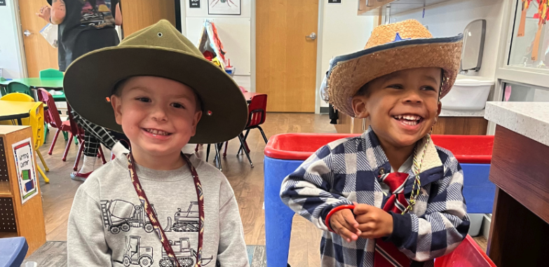 Two preschool boys in a preschool classroom wearing cowboy hats and very big smiles. They are playing dress up in the classroom.